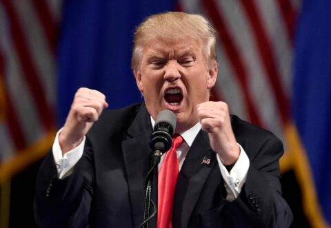 Blonde man in suit speaks passionately at Trump-style rally.